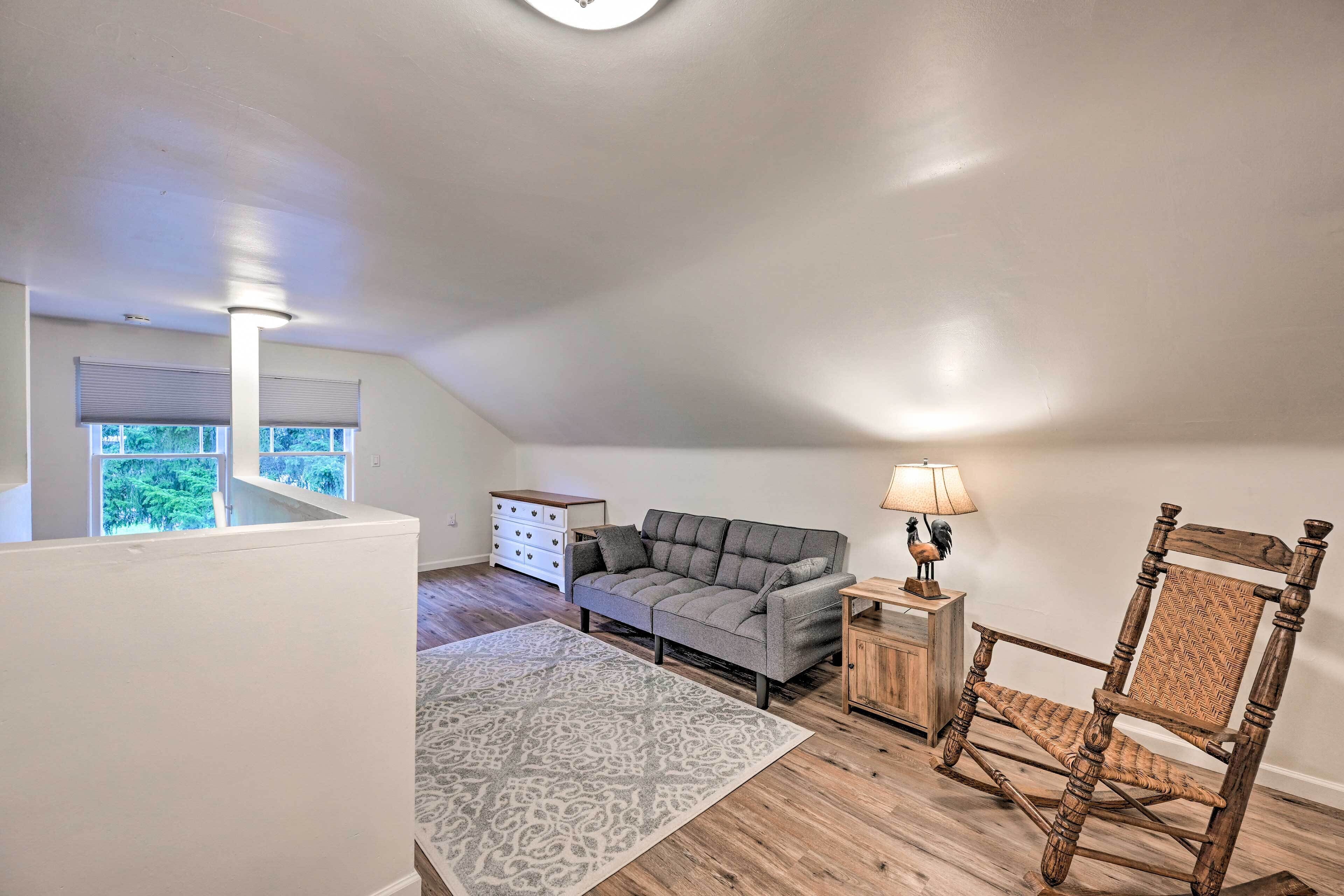 A loft-style room with a gray sofa, a wooden rocking chair, a patterned rug, and a small wooden side table with a lamp.