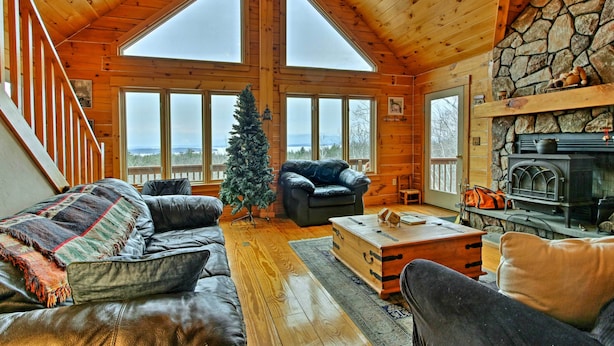 A cozy cabin interior with a stone fireplace, a Christmas tree, and a large window overlooking a snowy landscape. - Spacious Gilford Retreat w/ Deck: 2 Mi to Skiing! (Gilford)