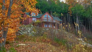 A large log cabin with a stone foundation, surrounded by autumn foliage and a forest backdrop. - Spacious Gilford Retreat w/ Deck: 2 Mi to Skiing! (Gilford)
