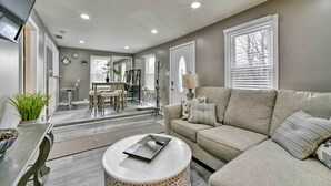 A modern living room with a beige sofa, a round coffee table, and a dining area with a table and chairs. - Completely Renovated Escape ~ 1 Mile to Downtown! (Farmington Hills)