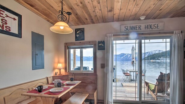 A cabin interior with a wooden bench, table, and a view of a lake through large glass doors. - Serenity at Scenic Bay: Floating Cottage w/ Views! (Bayview)