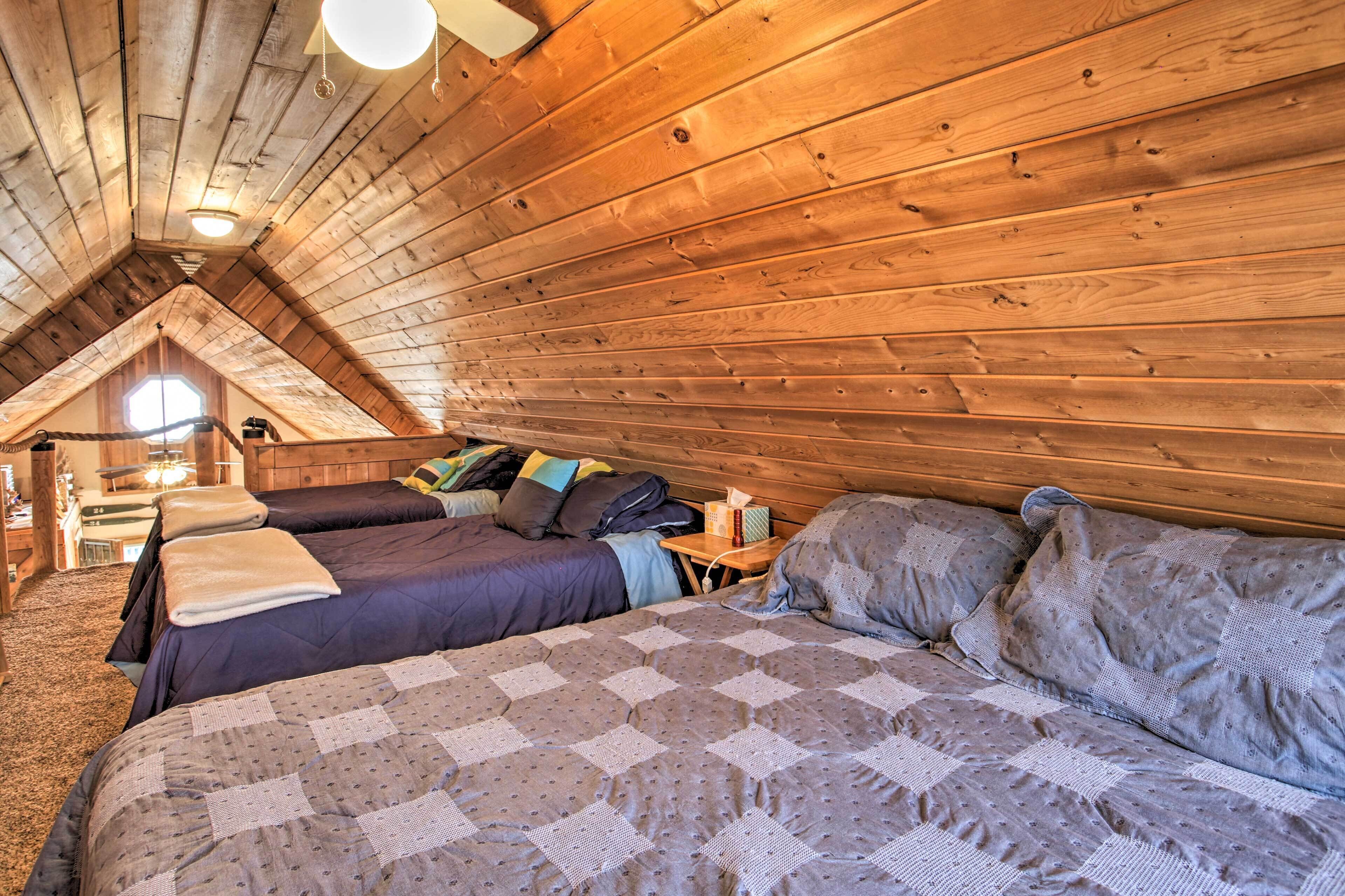 A cozy attic bedroom with wooden walls and a sloped ceiling, featuring two beds with blue and gray bedding, a small table, and a window with a view of the outside.