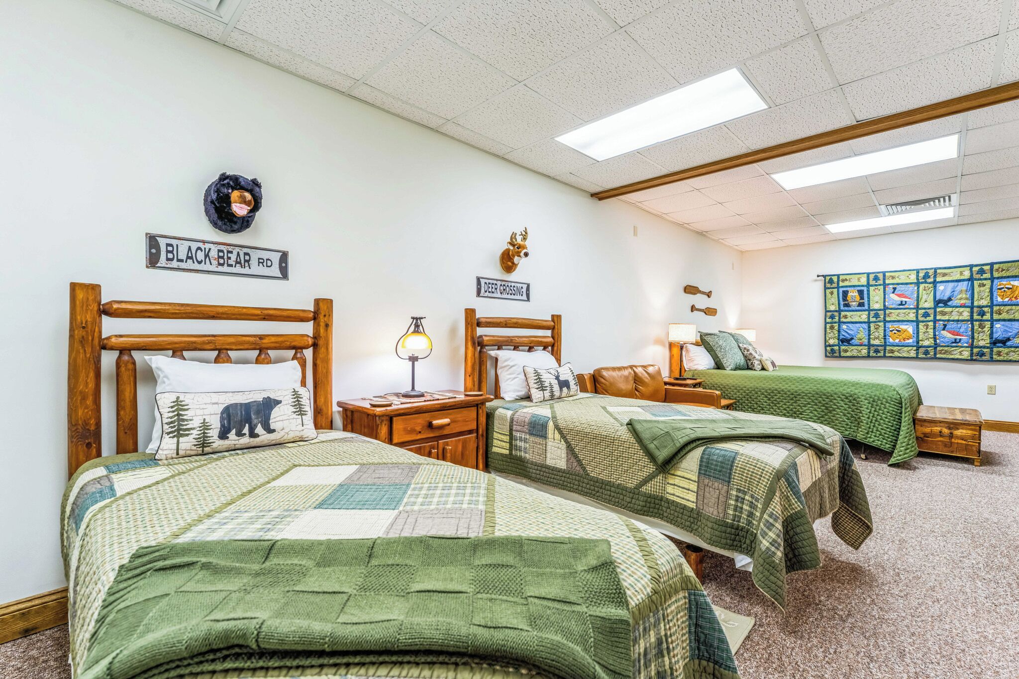 A room with two beds, each with a wooden headboard, a nightstand, a lamp, and a pillow with a bear design. There are wall decorations including a deer head and a quilted wall hanging.