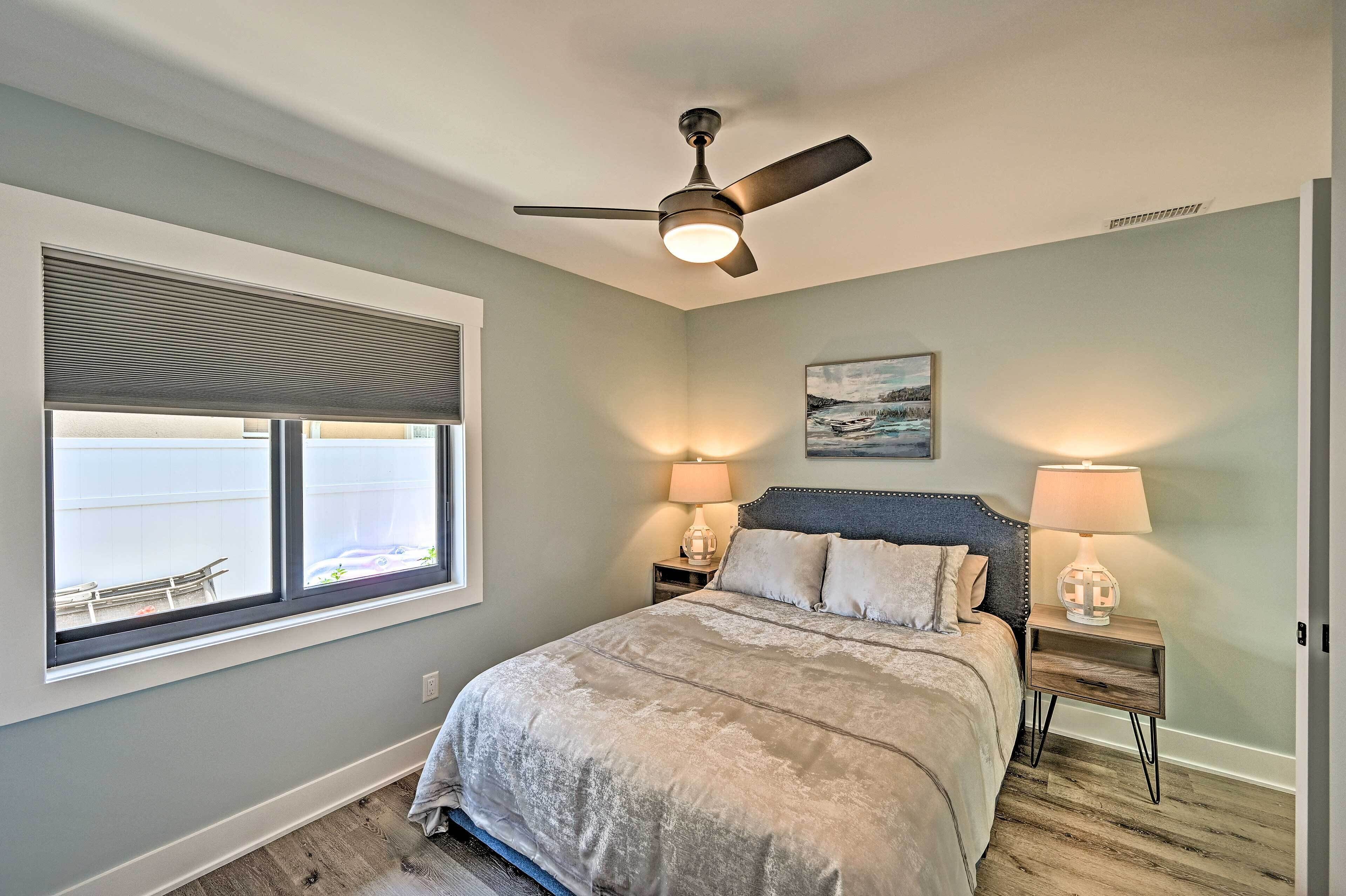 A bedroom with a bed, two bedside tables with lamps, a ceiling fan, a window with blinds, and a framed picture on the wall.