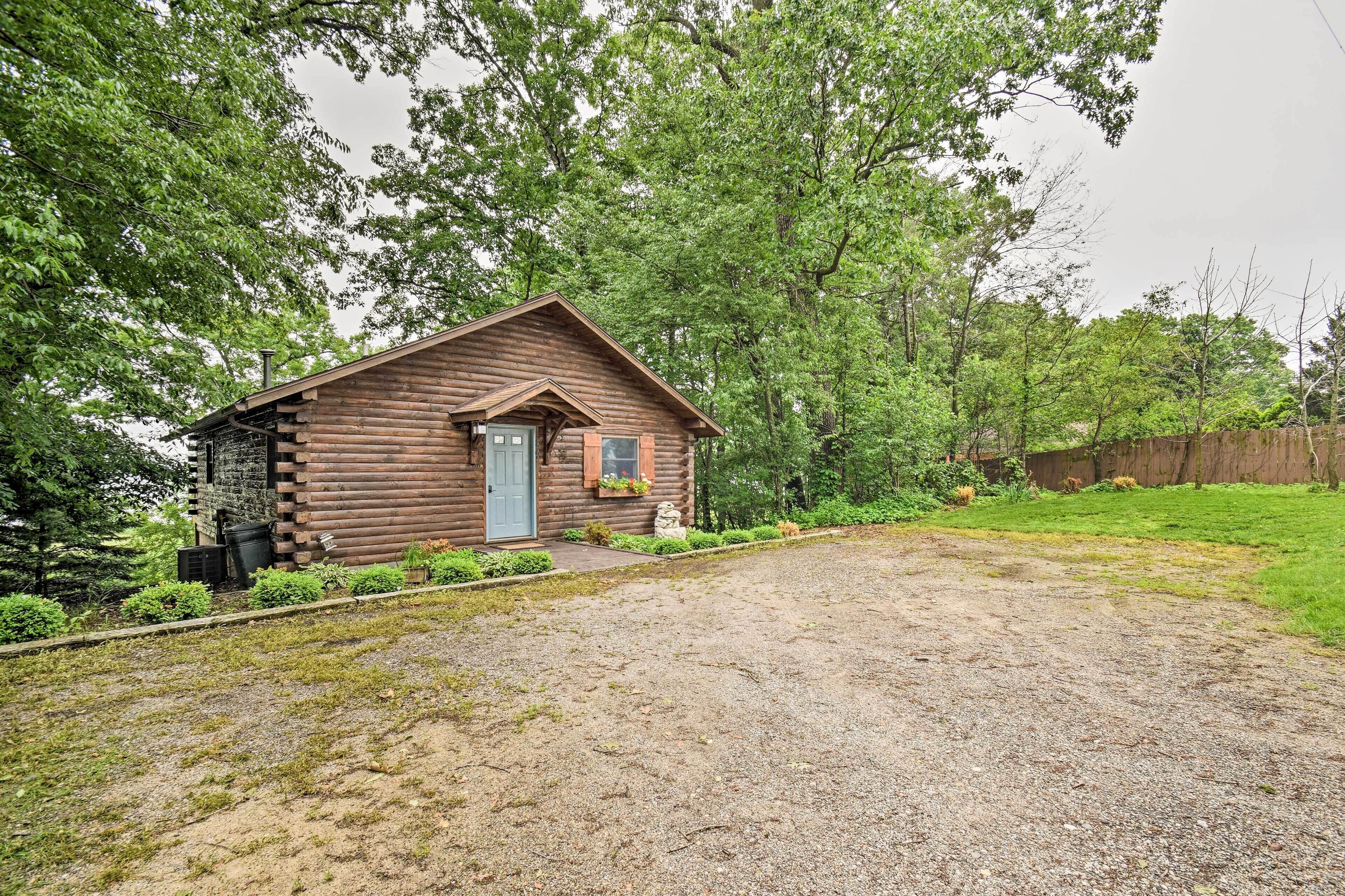 Une cabane rustique en rondins de bois, avec une porte d’entrée, un petit porche doté d’un banc et une allée gravillonnée.