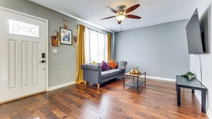 Sala de estar con piso de madera, un sofá gris con cojines coloridos, un ventilador de techo, una mesita con una planta y un televisor instalado en la pared.