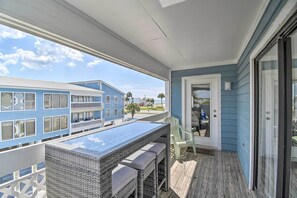 A balcony with a glass-top table and wicker chairs, overlooking a residential area with blue buildings. - Gulf Shores Condo: Private Balcony & Beach Views! (Gulf Shores)