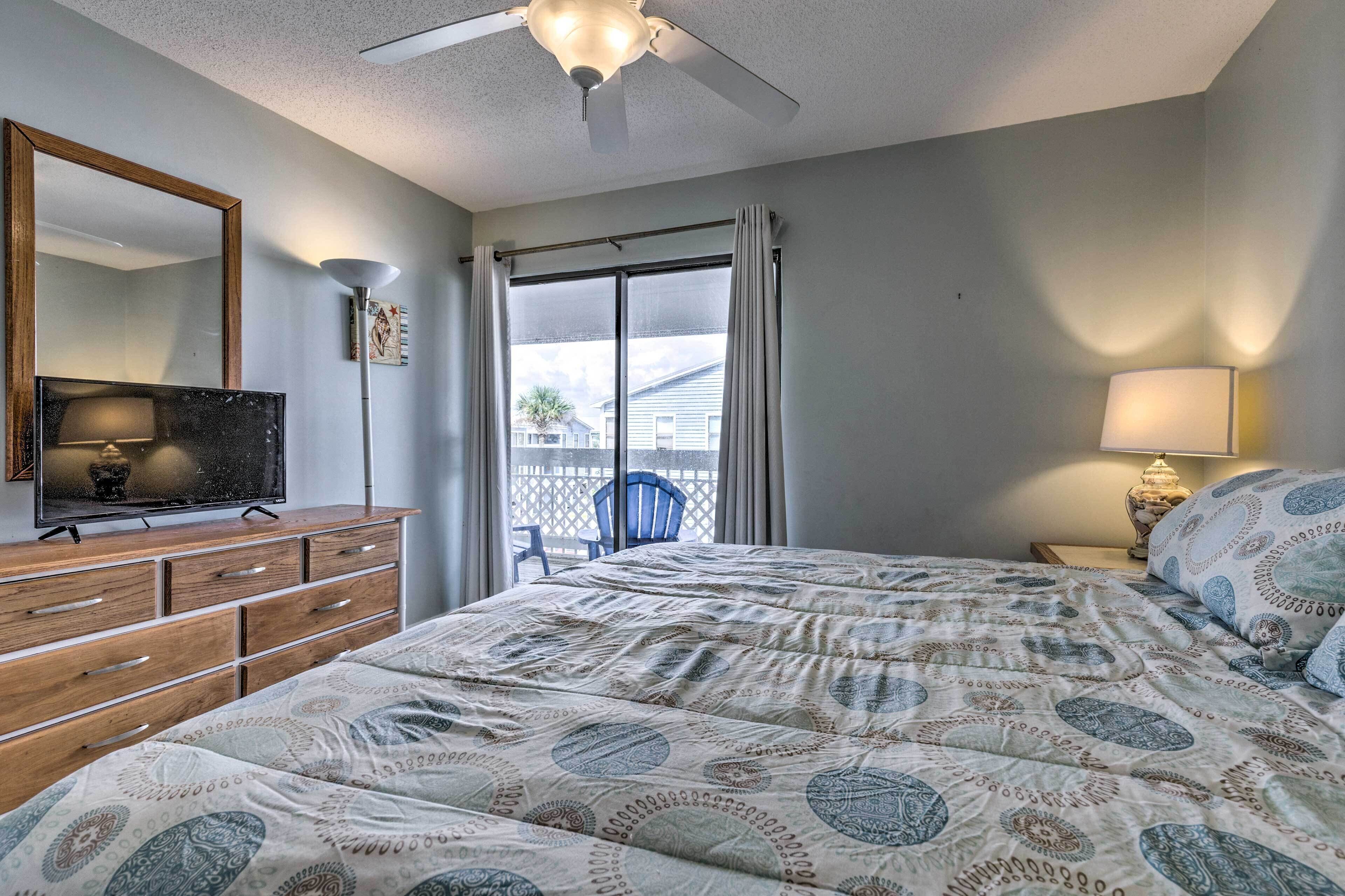 Une chambre à coucher avec un lit, un téléviseur posé sur une commode en bois, un ventilateur de plafond, une fenêtre avec des rideaux et une table de chevet avec une lampe.