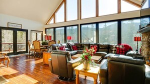 Ein geräumiges Wohnzimmer mit einer großen Eckcouch, einem Couchtisch und einem Kamin. Große Fenster bieten einen Blick ins Freie.