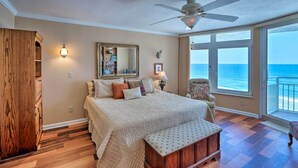 A bedroom with a large bed, a ceiling fan, a wooden wardrobe, a chair, and a view of the ocean.