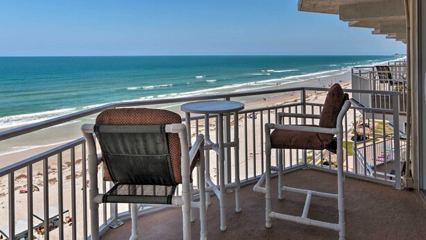 A balcony with two chairs and a small table overlooking a beach with waves and a clear sky.