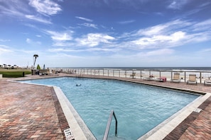En rektangulär swimmingpool med en terrass av tegelsten, ett metallräcke och utsikt över havet.