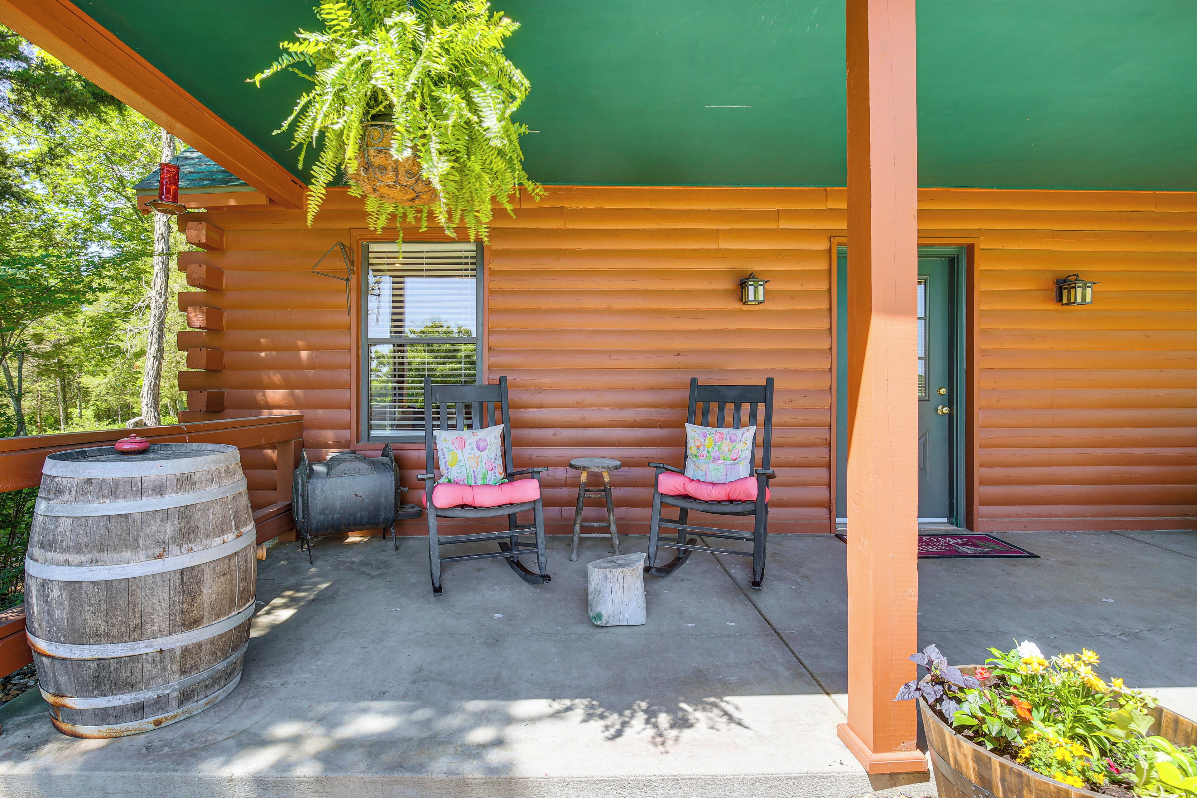 A wooden cabin with a porch, two rocking chairs, a small table, a barrel, and a planter with flowers.