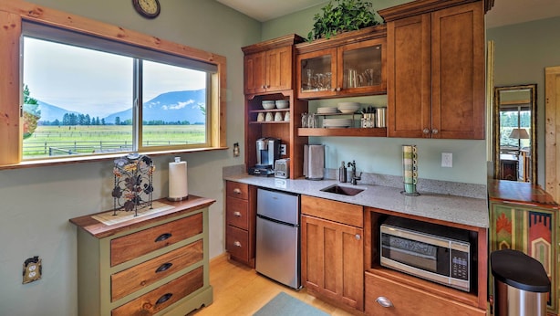 A kitchen with wooden cabinets, a stainless steel refrigerator, a microwave, and a window with a view of mountains. - Cabin w/ Porch & View ~ 19 Mi to West Glacier (Columbia Falls)