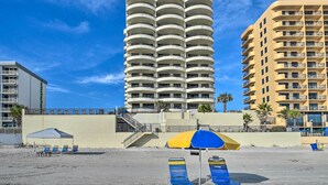 Un hôtel en bord de mer doté de nombreux balcons, d’une plage de sable avec des chaises longues et d’un ciel d’un bleu éclatant.
