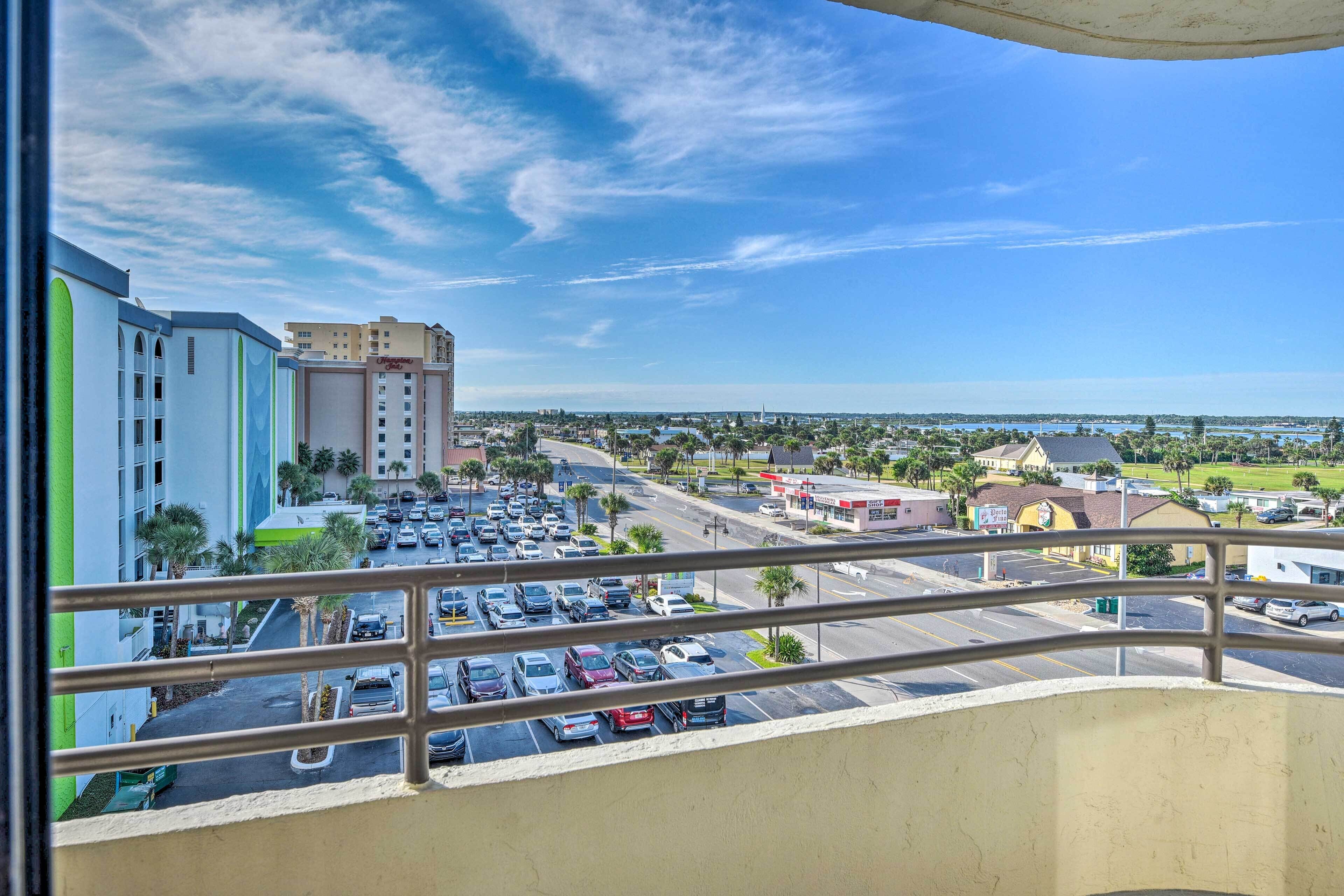 A view from a balcony overlooking a parking lot with several buildings in the background, including a hotel and a gas station.