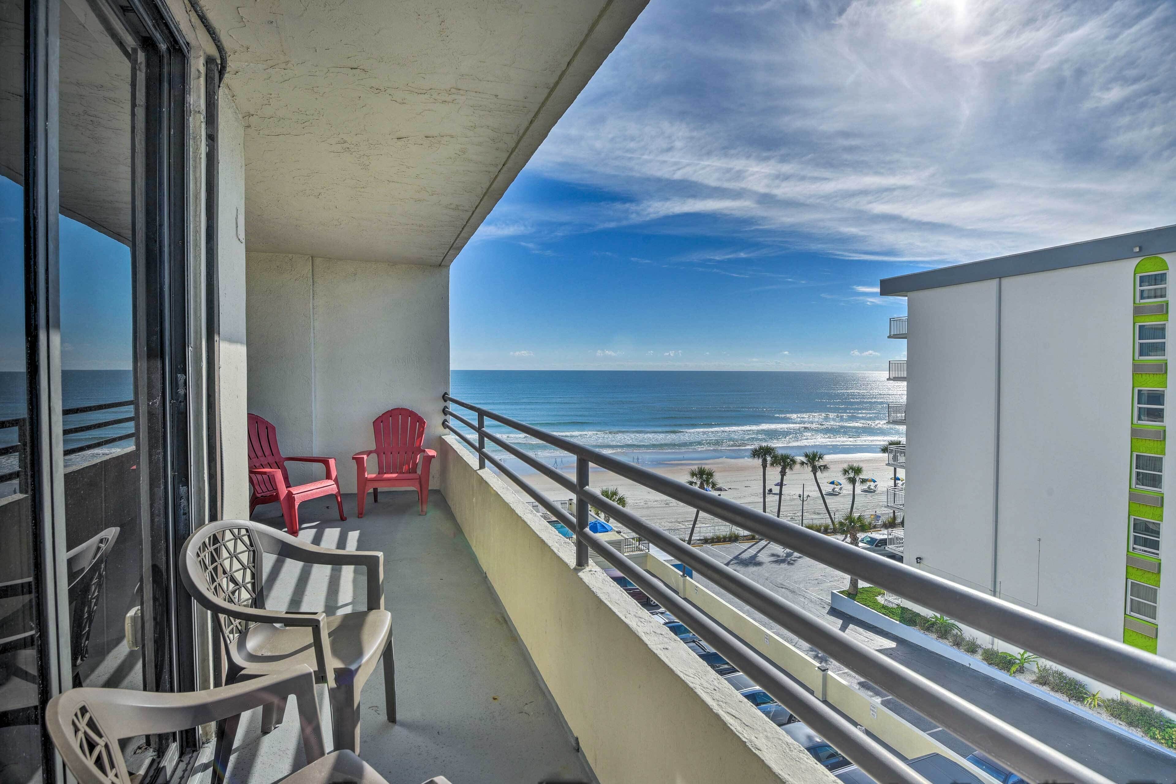 A balcony with a view of the beach, red chairs, and a white building with a green window.