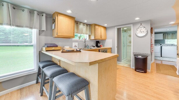 A modern kitchen with wooden cabinets, a central island with bar stools, a clock, and a view of the outside through a window with blinds. - Visit Fife Lake & Traverse Bay! Farmhouse Hideaway (Fife Lake)