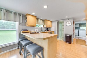 A modern kitchen with wooden cabinets, a central island with bar stools, a clock, and a view of the outside through a window with blinds. - Visit Fife Lake & Traverse Bay! Farmhouse Hideaway (Fife Lake)