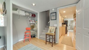 A compact, well-lit hallway with a wooden floor, a red chair, and a small seating area with a cushion. A staircase leads to an upper level with a closet containing folded clothes and a shelf with towels. - Visit Fife Lake & Traverse Bay! Farmhouse Hideaway (Fife Lake)
