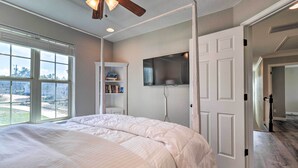 A bedroom with a large bed, a ceiling fan, a bookshelf, a flat-screen TV, and a view of the outdoors through a window. - Lake LBJ Waterfront Vacation Rental! (Marble Falls)