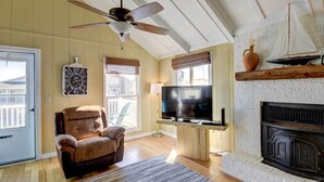 A cozy living room with a fireplace, a television, a ceiling fan, and a large window with blinds. - Ocean Isle Beach Home w/ Canal Views! (Ocean Isle Beach)