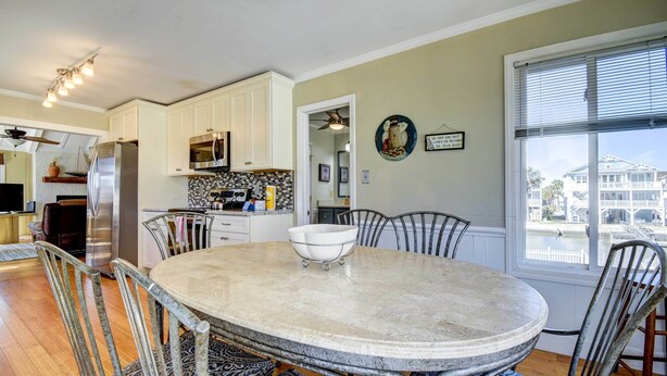 A dining area with a round table, chairs, and a view of a canal through a large window. - Ocean Isle Beach Home w/ Canal Views! (Ocean Isle Beach)