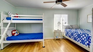 A room with two bunk beds, a ceiling fan, a window with blinds, and a small table with a lamp. - Ocean Isle Beach Home w/ Canal Views! (Ocean Isle Beach)