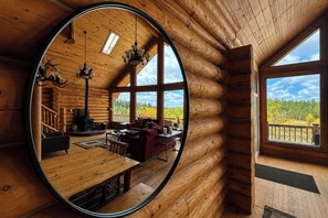 L’intérieur d’une cabane en rondins, avec un grand miroir qui reflète la pièce, une chemin ée, un coin salon confortable et une vue sur la forêt à travers de grandes fenêtres.