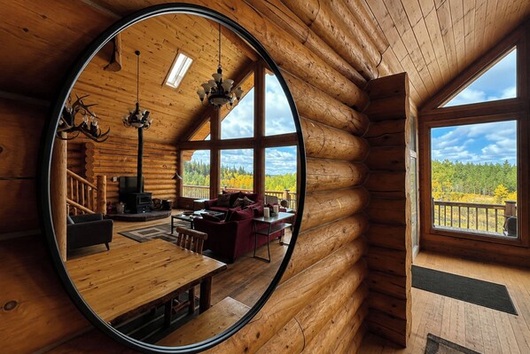 Interior de una cabaña de troncos con un espejo grande que refleja la habitación, una chimenea, una zona de estar acogedora y vistas al bosque a través de amplias ventanas.