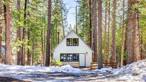 A cabin with a triangular roof, surrounded by tall pine trees, a wooden deck, and a snow-covered ground.