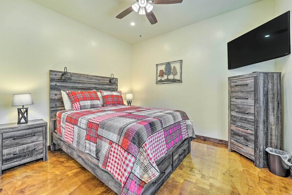 A bedroom with a wooden headboard, a bed with a red and gray quilt, a nightstand with a lamp, a dresser, and a television on the wall.