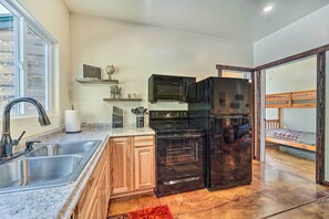 A compact kitchen with a sink, stove, microwave, and refrigerator. There is a wooden cabinet and a small shelf with kitchen utensils. A window provides natural light, and a red rug is on the floor. - Serene Dolores Cabin w/ Patio & River Access! (Dolores)