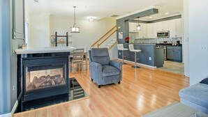Ein modernes Wohnzimmer mit Kamin, einem grauen Sessel, Holzböden und einer Küche im Hintergrund.