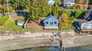 Vue aérienne d’une propriété située sur la côte, avec une maison, un petit abri et une jetée s’étendant dans une eau d’un bleu limpide.