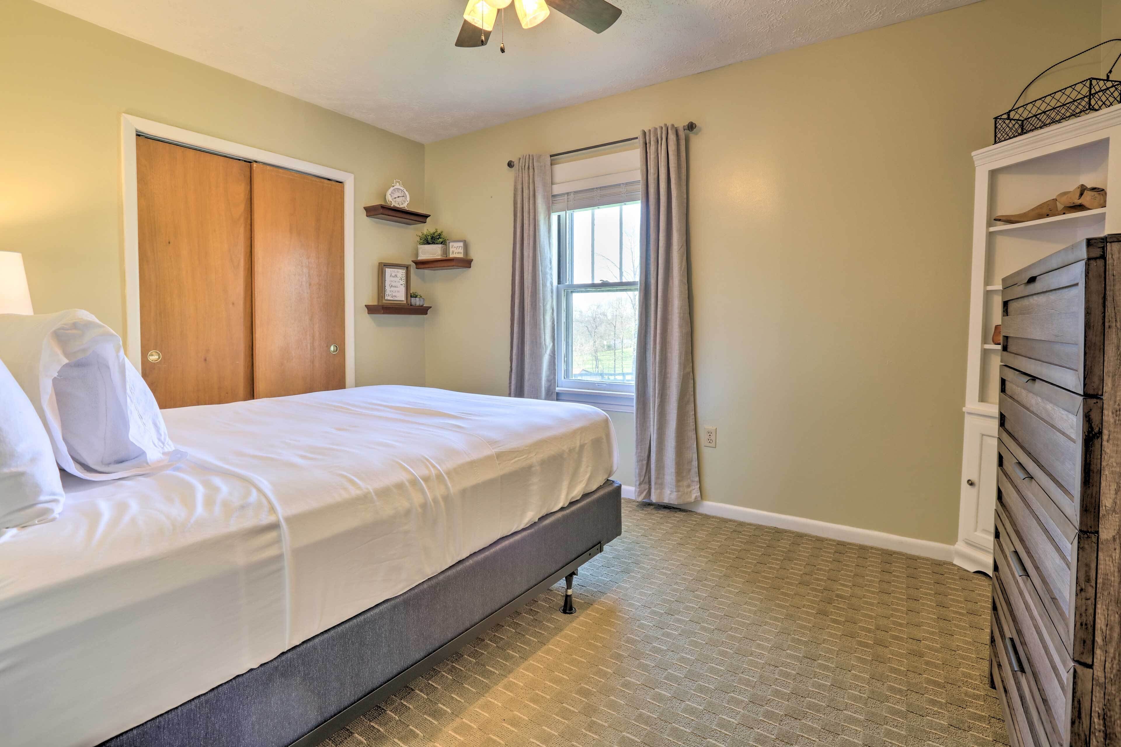 A neatly made bed in a room with a wooden wardrobe, a window with curtains, and a shelf with decorative items.