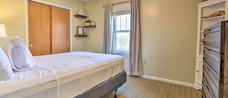 A neatly made bed in a room with a wooden wardrobe, a window with curtains, and a shelf with decorative items.