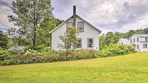 A white house with a gabled roof and a chimney, surrounded by lush greenery and a well-maintained lawn. - Pet-friendly Roxbury Farmhouse on 350 Acres! (Roxbury)