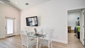 Un área de comedor con una mesa de vidrio y sillas blancas, un televisor de pantalla plana montado en la pared, y un cuarto de lavandería con lavadoras.