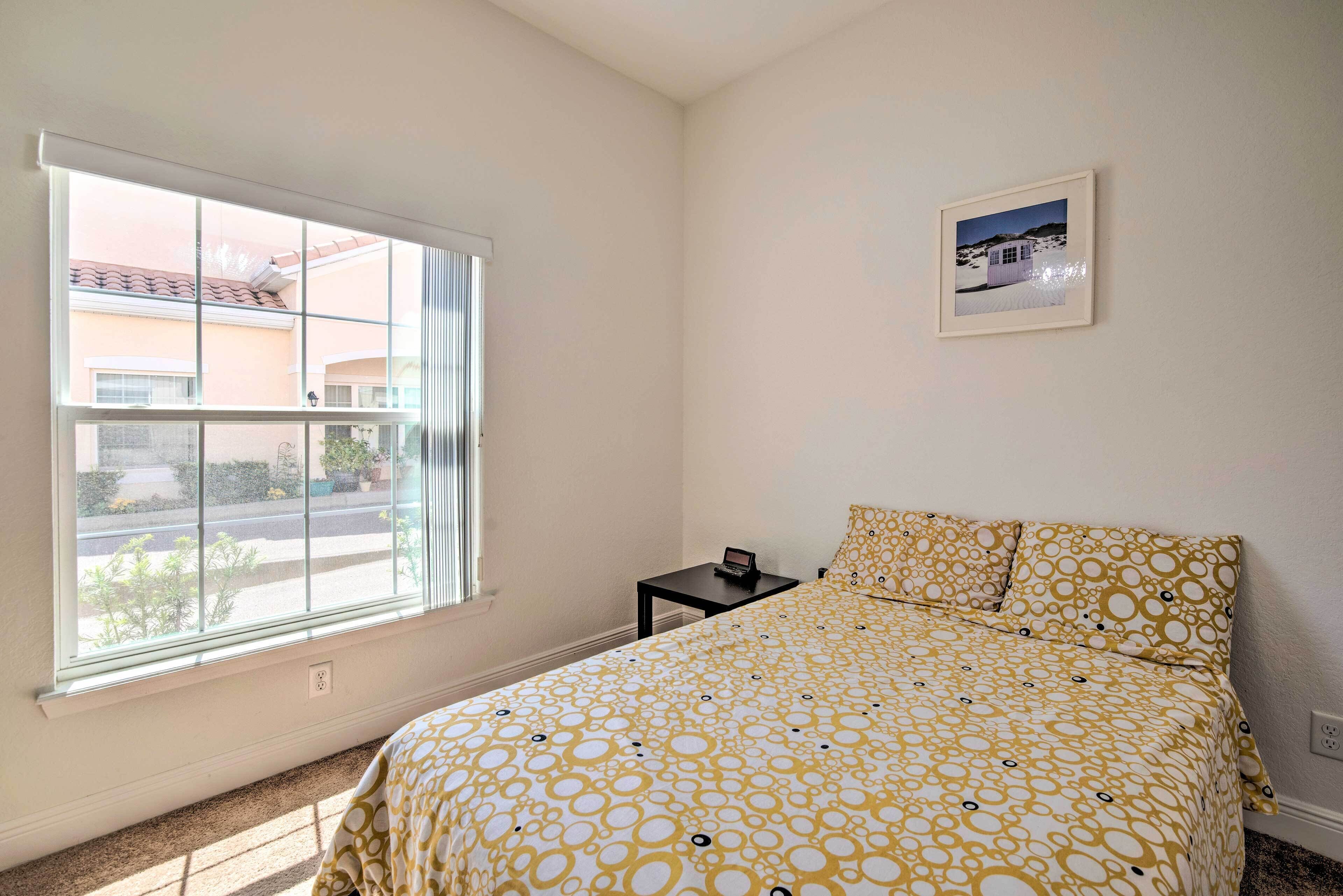 A bedroom with a large window, a bed with patterned bedding, a small bedside table, and a framed picture on the wall.