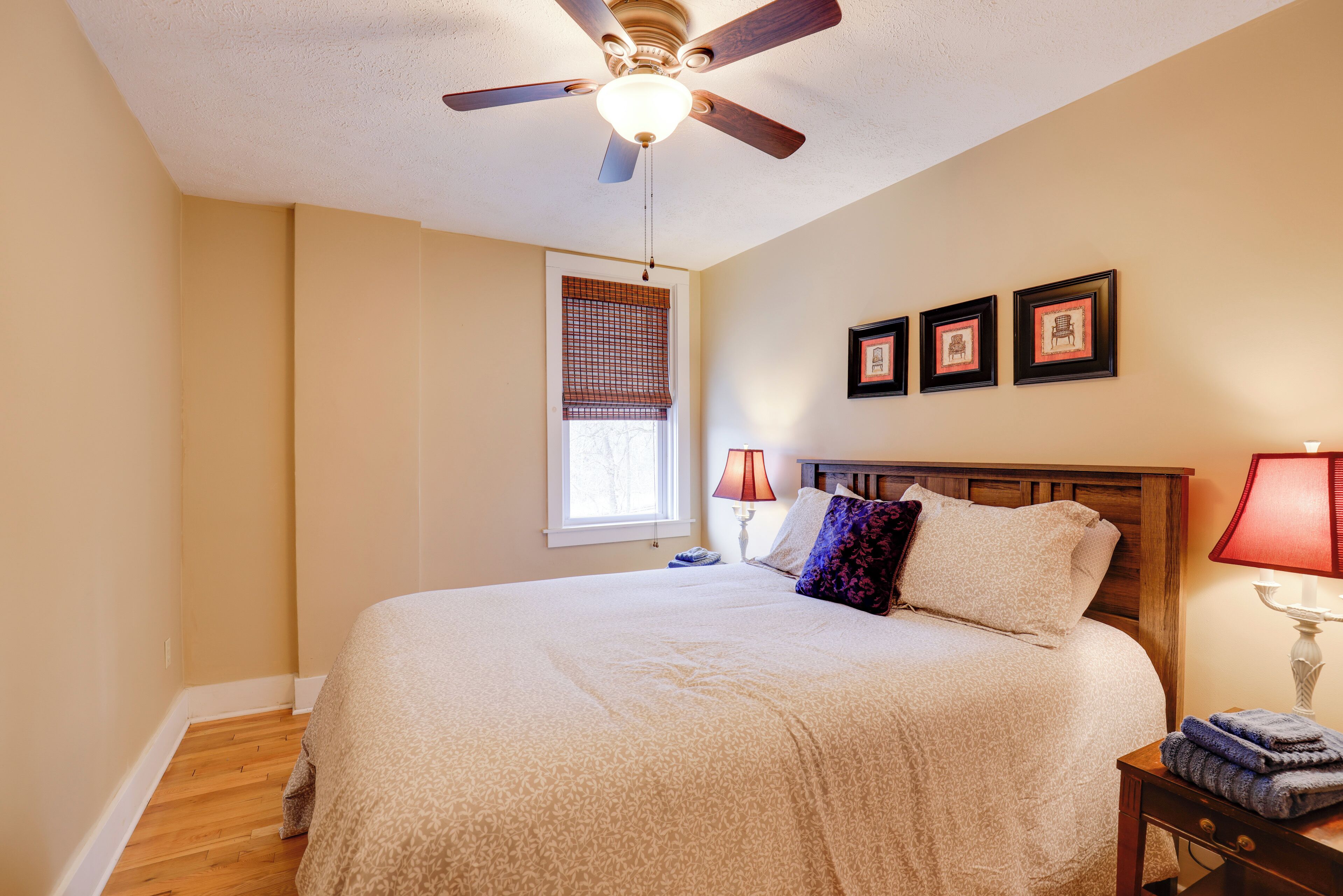 A bedroom with a bed, a ceiling fan, a window with blinds, a nightstand with a lamp, and framed pictures on the wall.