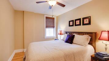 A bedroom with a bed, a ceiling fan, a window with blinds, a nightstand with a lamp, and framed pictures on the wall.