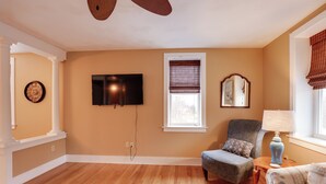 A room with a wooden floor, a flat-screen TV mounted on the wall, a window with blinds, a chair, a lamp, and a decorative mirror. - Finger Lakes Retreat: 3 Mi to Seneca & Keuka Lake (Himrod)
