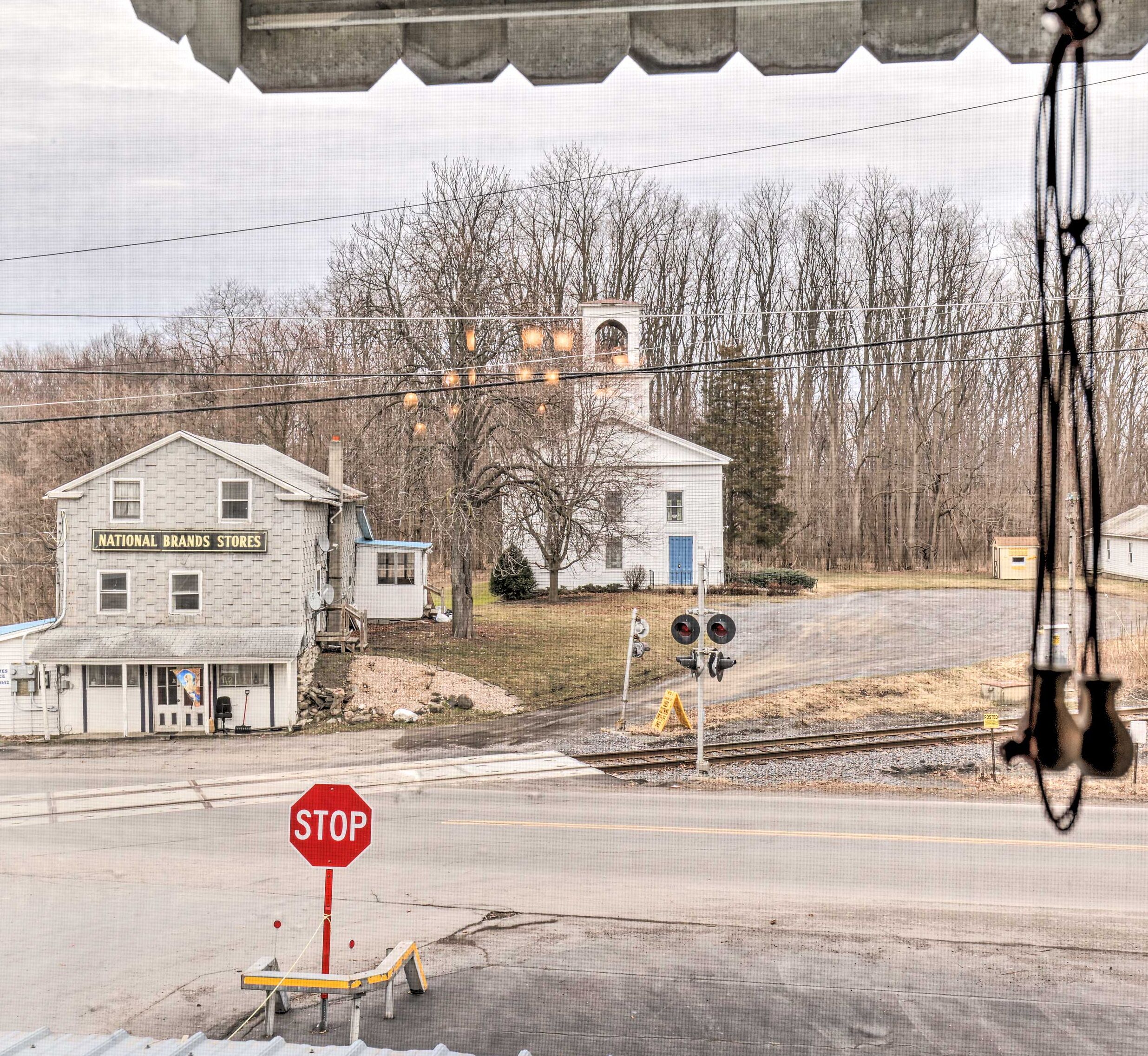 Une intersection située en zone rurale, avec un panneau de signalisation indiquant un arrêt obligatoire, un passage à niveau et un bâtiment portant l’inscription « National Brands Stores ».