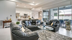 A modern living room with leather sofas, a glass coffee table, and a view of the kitchen and outdoor patio.