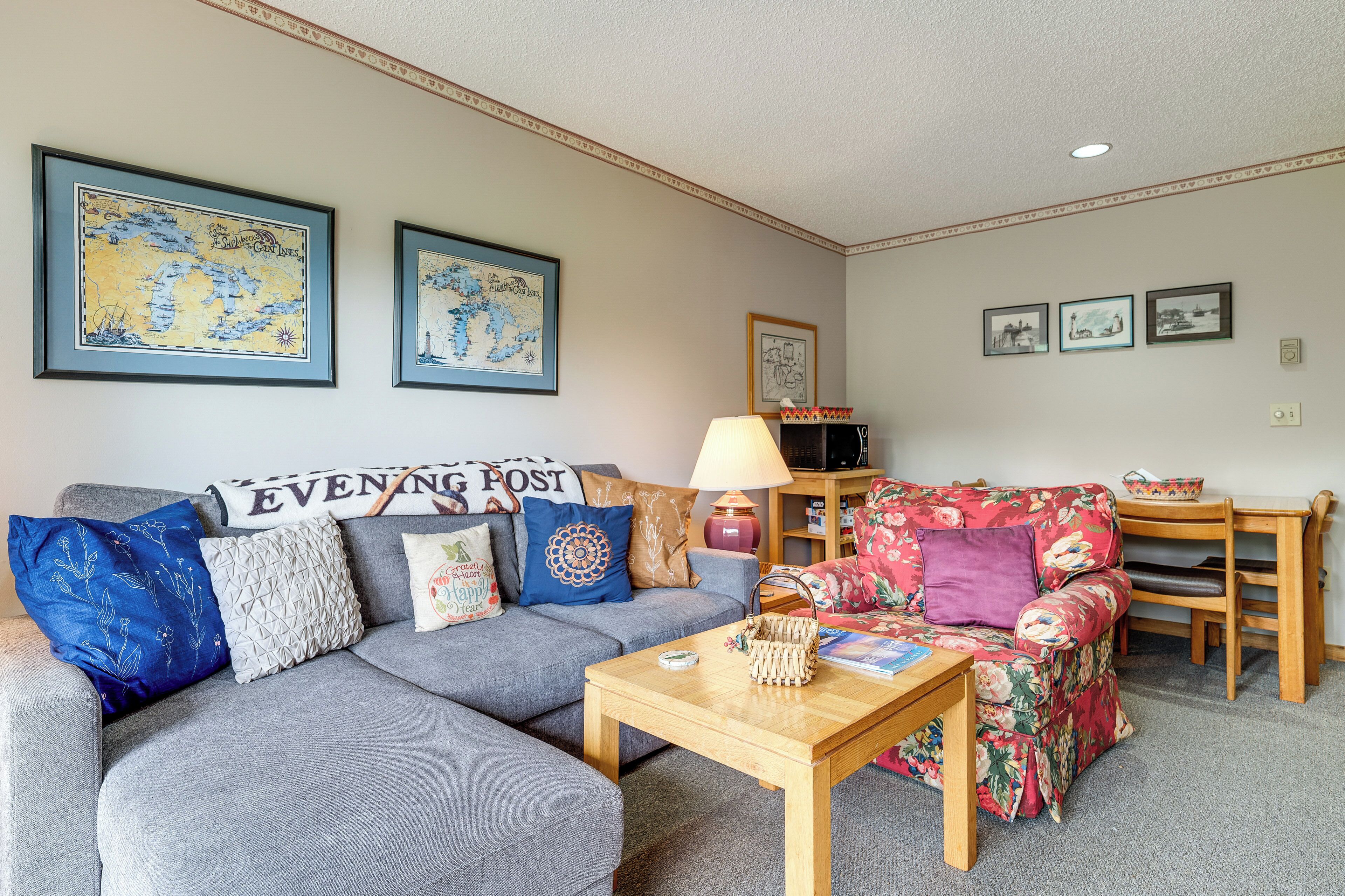A living room with a grey sofa, a floral armchair, a wooden coffee table, and a lamp. There are framed pictures on the wall and a sign that reads 'EVENING POST'.
