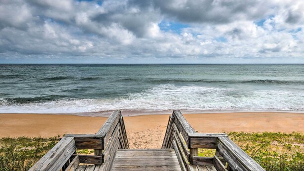 A wooden boardwalk leading to a beach with waves crashing onto the shore under a cloudy sky. - Sun-soaked Condo w/ Balcony - Steps to Beach! (Ormond Beach)