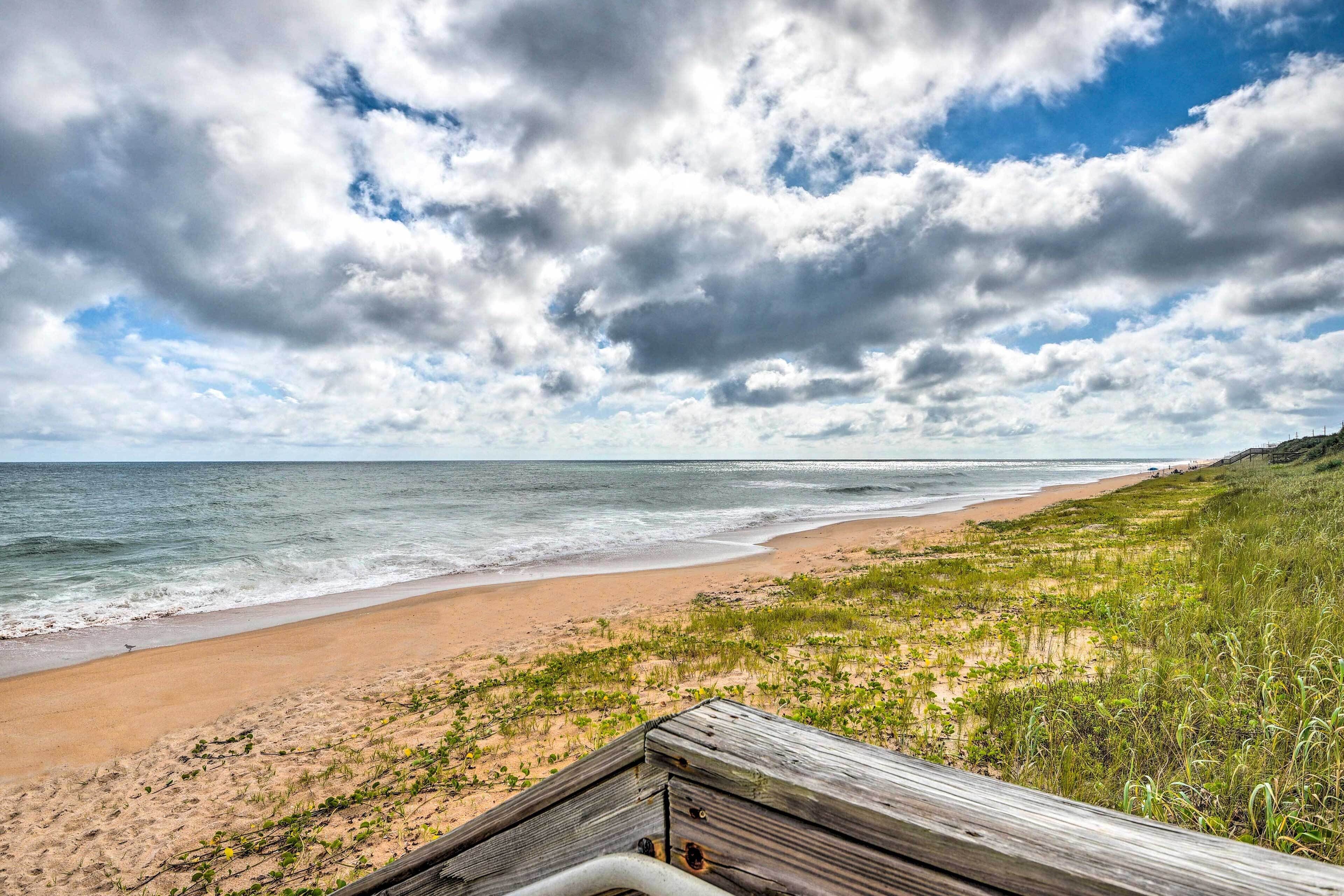 Ein Strand mit einer hölzernen Promenade, die zum Ufer führt, grasbewachsenen Dünen und einem teilweise bewölkten Himmel.