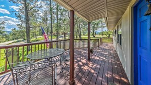 Eine überdachte Veranda mit Tisch und Stühlen, eine blaue Tür sowie Blick auf Bäume und eine Flagge.
