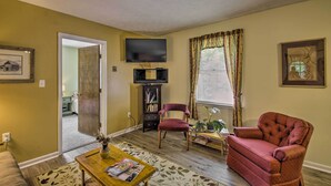 A living room with a TV, a red armchair, a bookshelf, and a window with curtains. - Quaint Hinton Hideaway - Hunt & Fish Nearby! (Hinton)